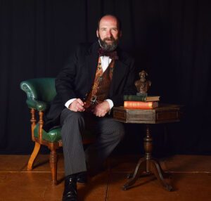 Gerald Dickens, sitting at a desk, in period costume